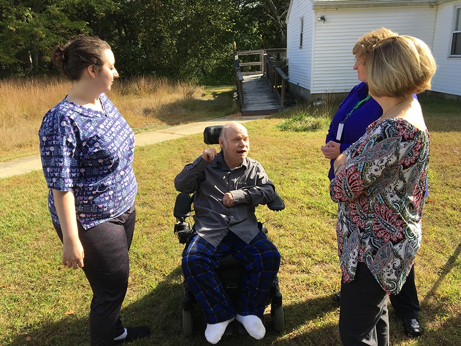 A man in a wheelchair standing with caregivers and neighbor in the yard