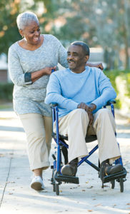 A senior African American couple taking a walk. The man is sitting in a wheelchair being pushed by his wife. They are talking and smiling.